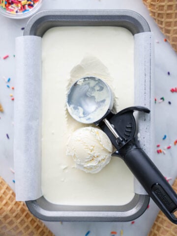 No Churn Ice Cream in a loaf pan with an ice cream scooper and sprinkles and waffle cone nearby.