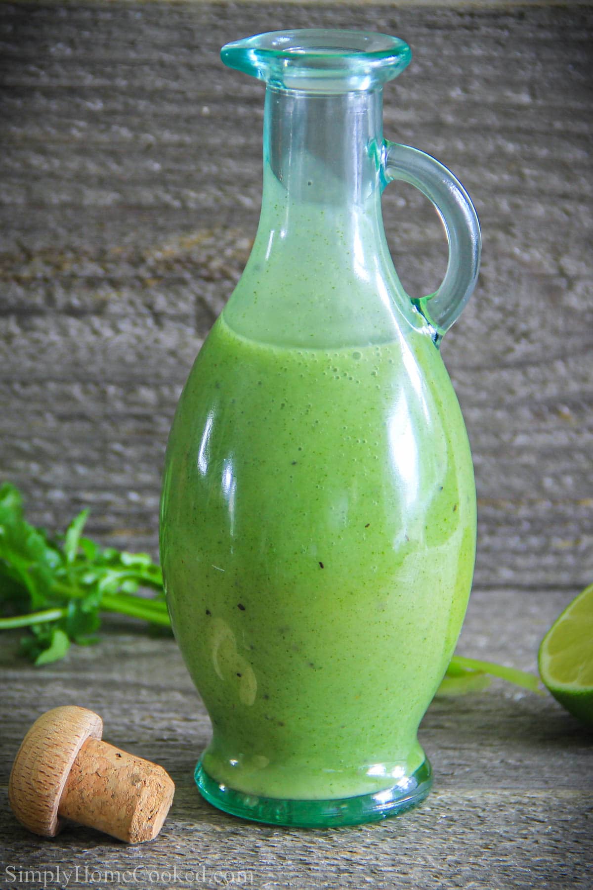 This Easy Cilantro Lime Dressing is a mix of sweet, sour, and tart. You'll love the way the flavors come together and go with your salad. #easycilantrolimedressing #easydressingrecipe #cilantrolimedressing #cilantrolimedressingrecipe Cilantro Lime dressing in a carafe with a stopper, lime, and cilantro in the background
