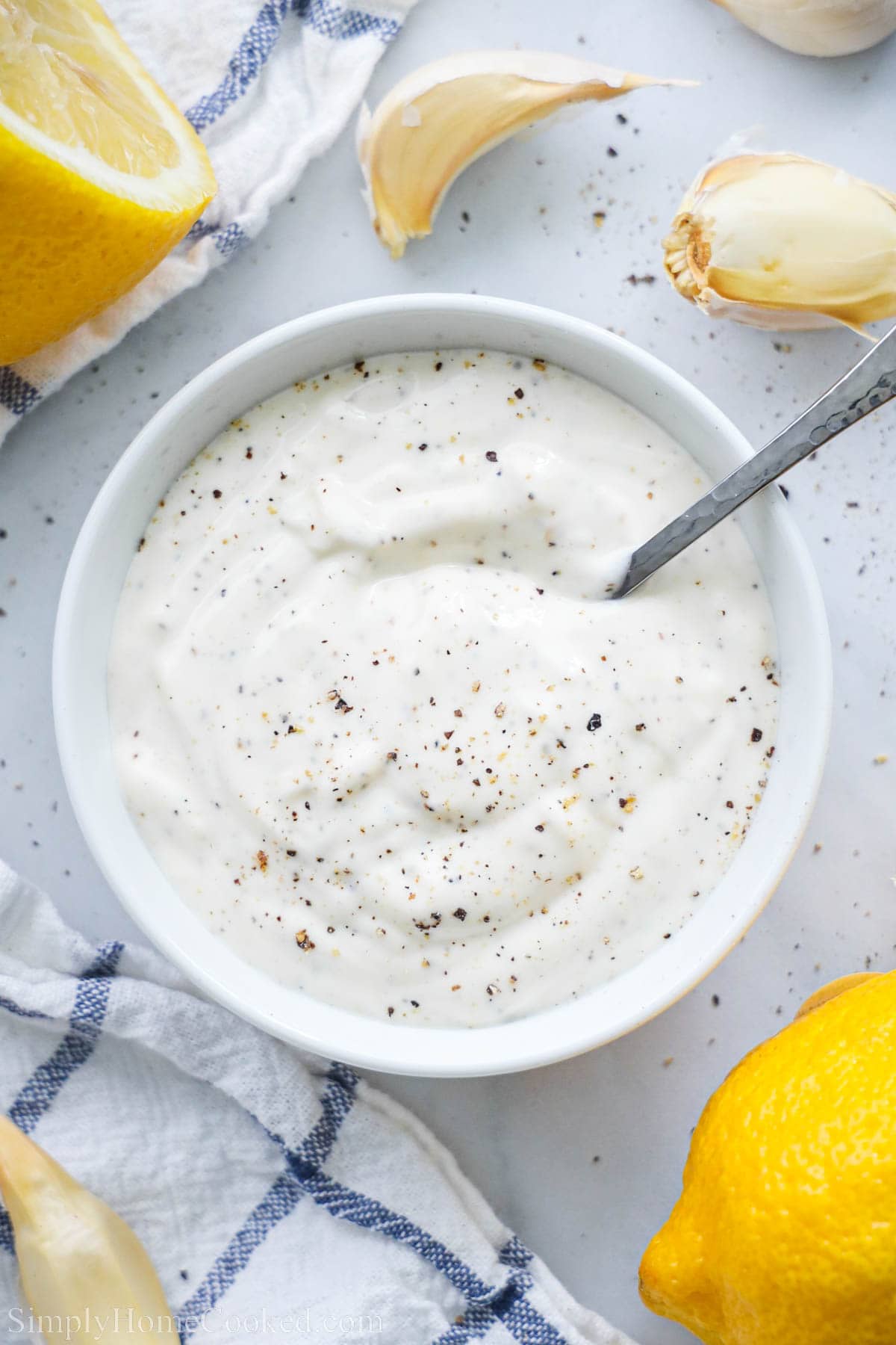 Garlic Aioli in a bowl with a spoon and lemons and garlic cloves nearby.