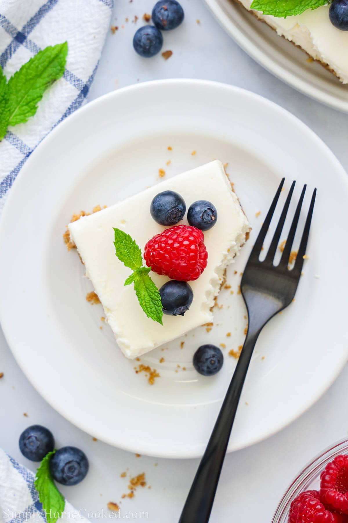 Overview of No-bake Cheesecake Bar missing a bite on a plate and topped with blueberries and a raspberry, with a fork nearby.