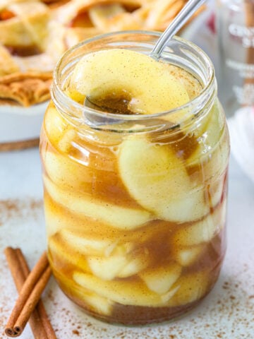 Homemade Apple Pie Filling in a mason jar with a spoon and cinnamon sticks nearby.