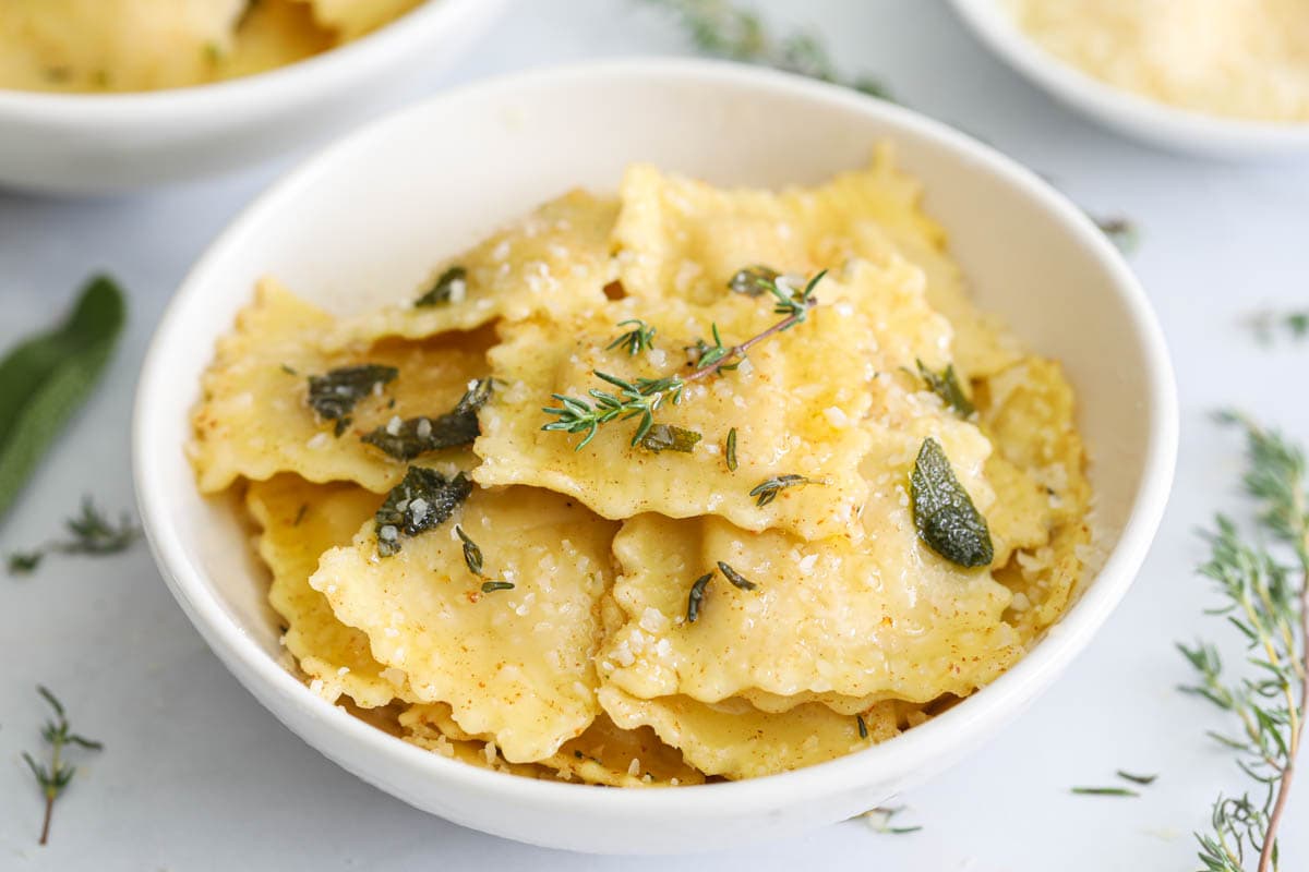 This Butternut Squash Ravioli is a fresh and delicious fall dish that you are going to love. Made with fresh herbs, Parmesan, and brown butter, the flavors of this pasta are absolutely mouthwatering. #raviolirecipe #homemaderavioli #butternutsquashraviolirecipe #fallbutternutsquashravioli Butternut Squash Ravioli topped with fresh thyme, sage, and Parmesan cheese.