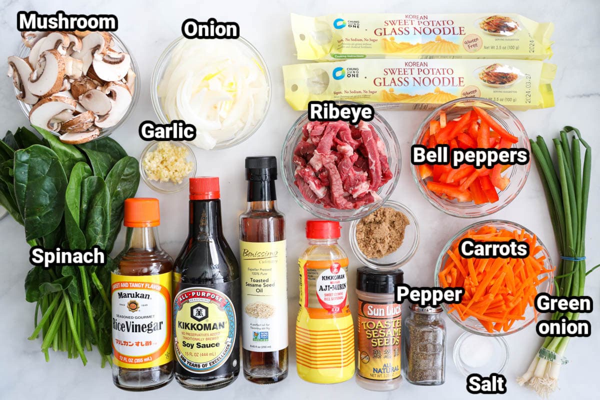 Ingredients in Easy Japchae Recipe: mushrooms, onion, garlic, spinach, glass noodles, ribeye, bell peppers, carrots, green onions, salt, pepper, brown sugar, toasted sesame seeds, aji mirin, toasted sesame oil, soy sauce, and ice vinegar. 