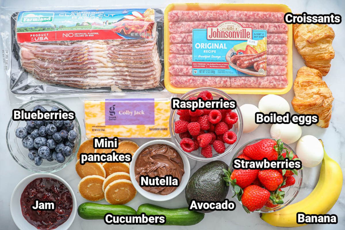 Ingredients for Breakfast Charcuterie Board: bacon, sausage links, croissants, blueberries, raspberries, strawberries, hard boiled eggs, banana, avocado, cucumber, Nutella, mini pancakes, colby jack cheese, and jam. 