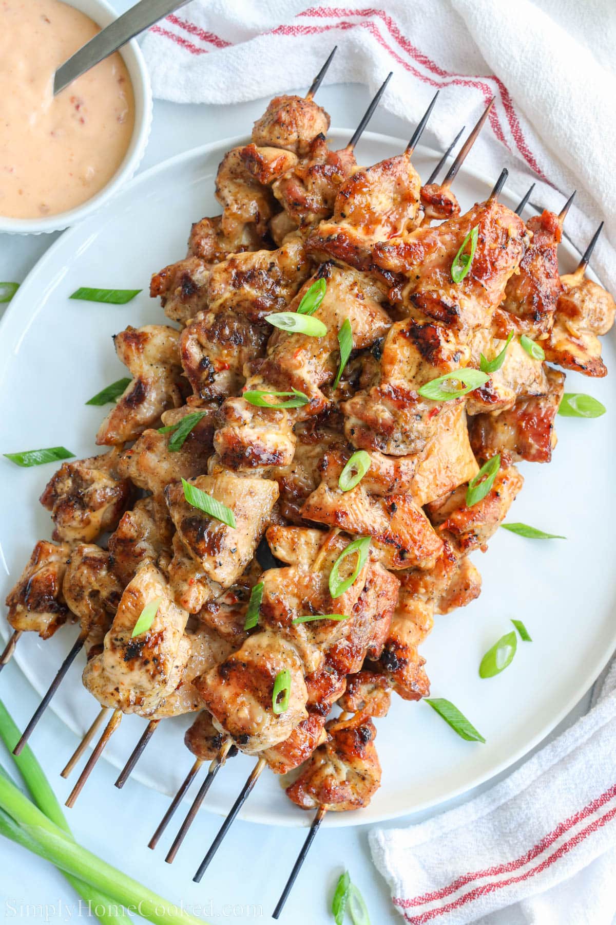 A pile of skewered bang bang chicken on a white plate with sliced green inion on top.