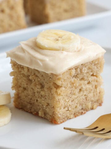 A slice of Banana Cake topped with brown butter cream cheese frosting and a slice of banana.