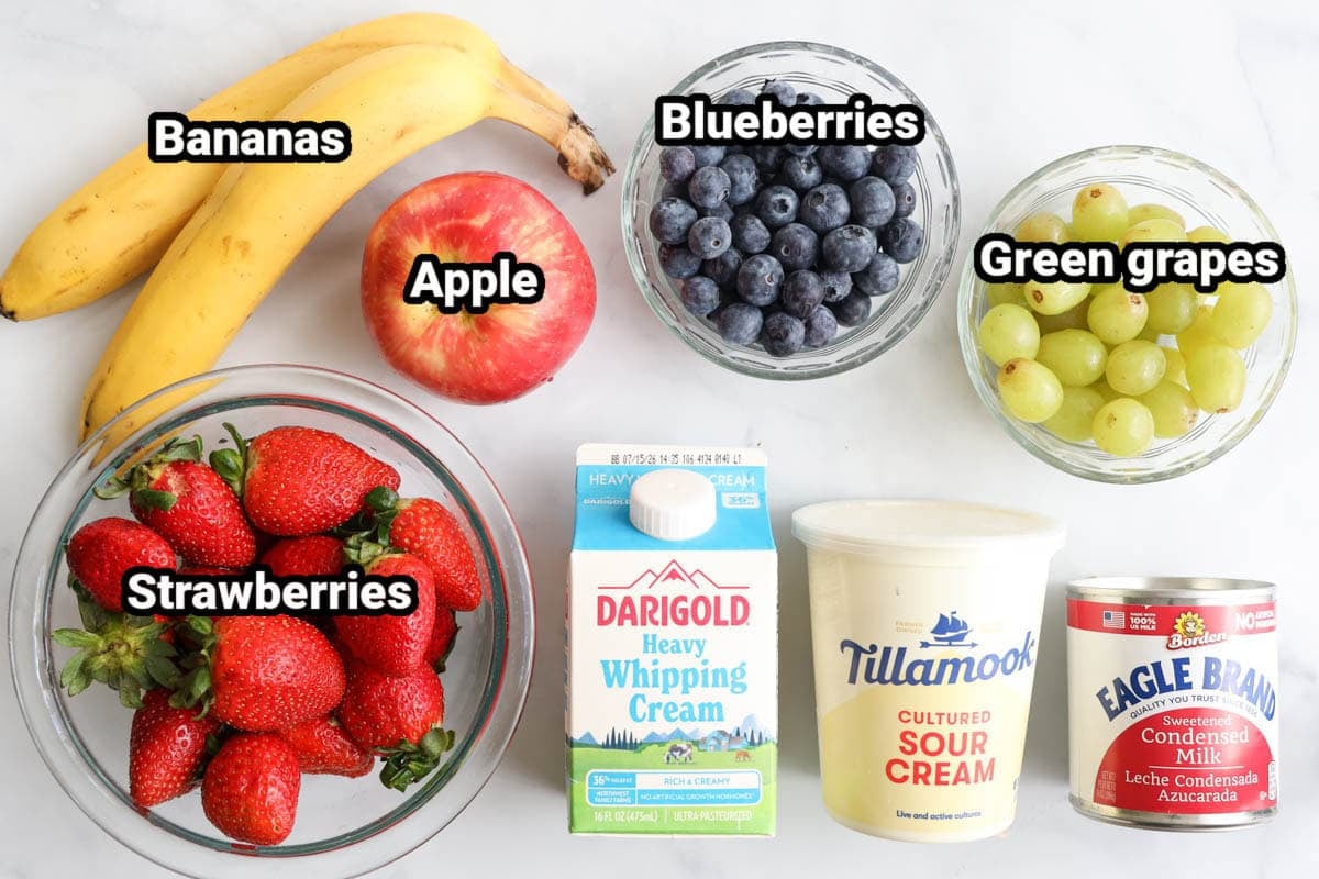 Ingredients for Creamy Fruit Salad: strawberries, blueberries, bananas, apple, green grapes, sour cream, heavy cream, and sweetened condensed milk.
