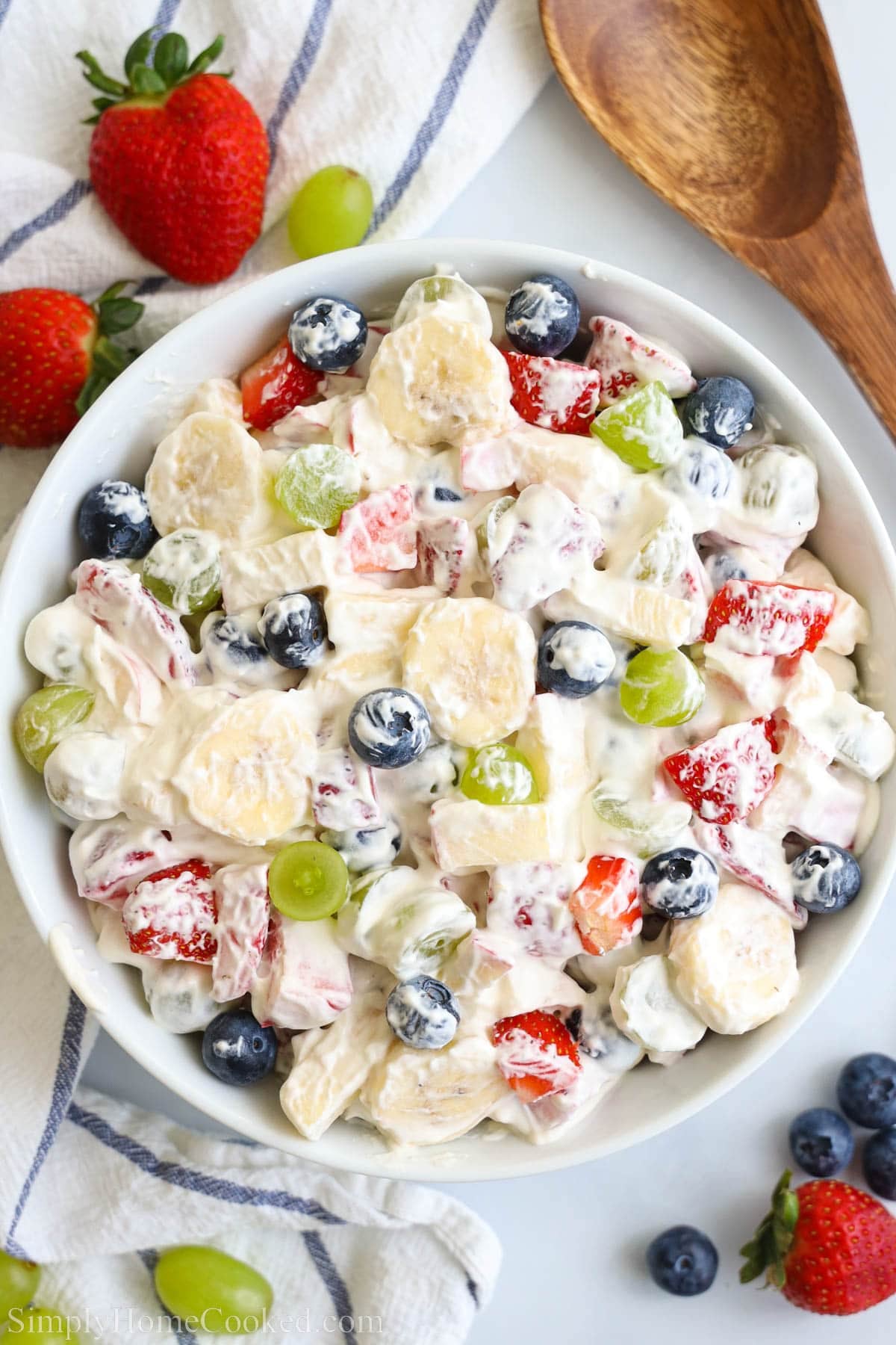 Overhead view of Creamy Fruit Salad in a bowl with fruit and a wooden spoon nearby.